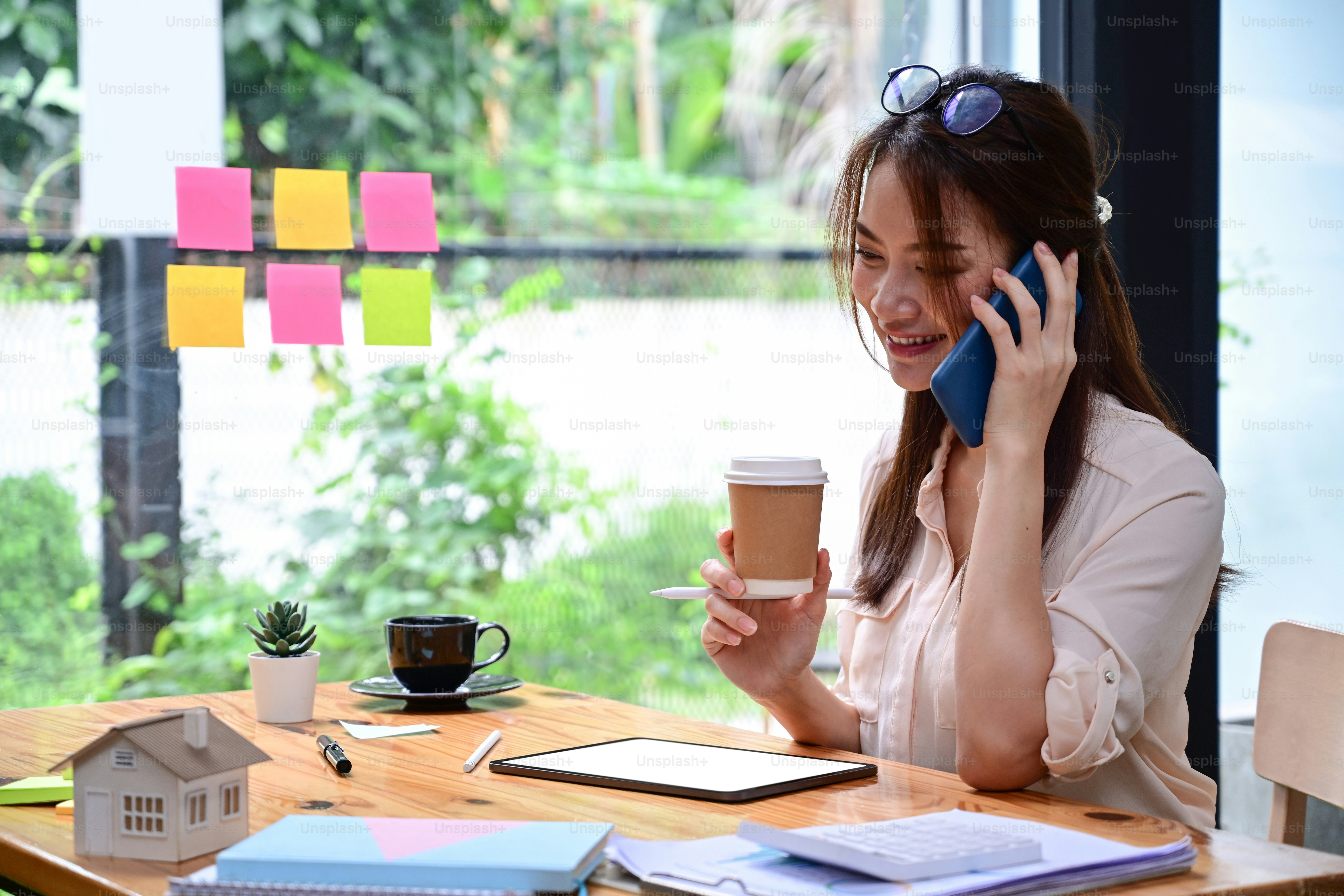 Smiling young female drinking coffee and talking on mobile phone at office.