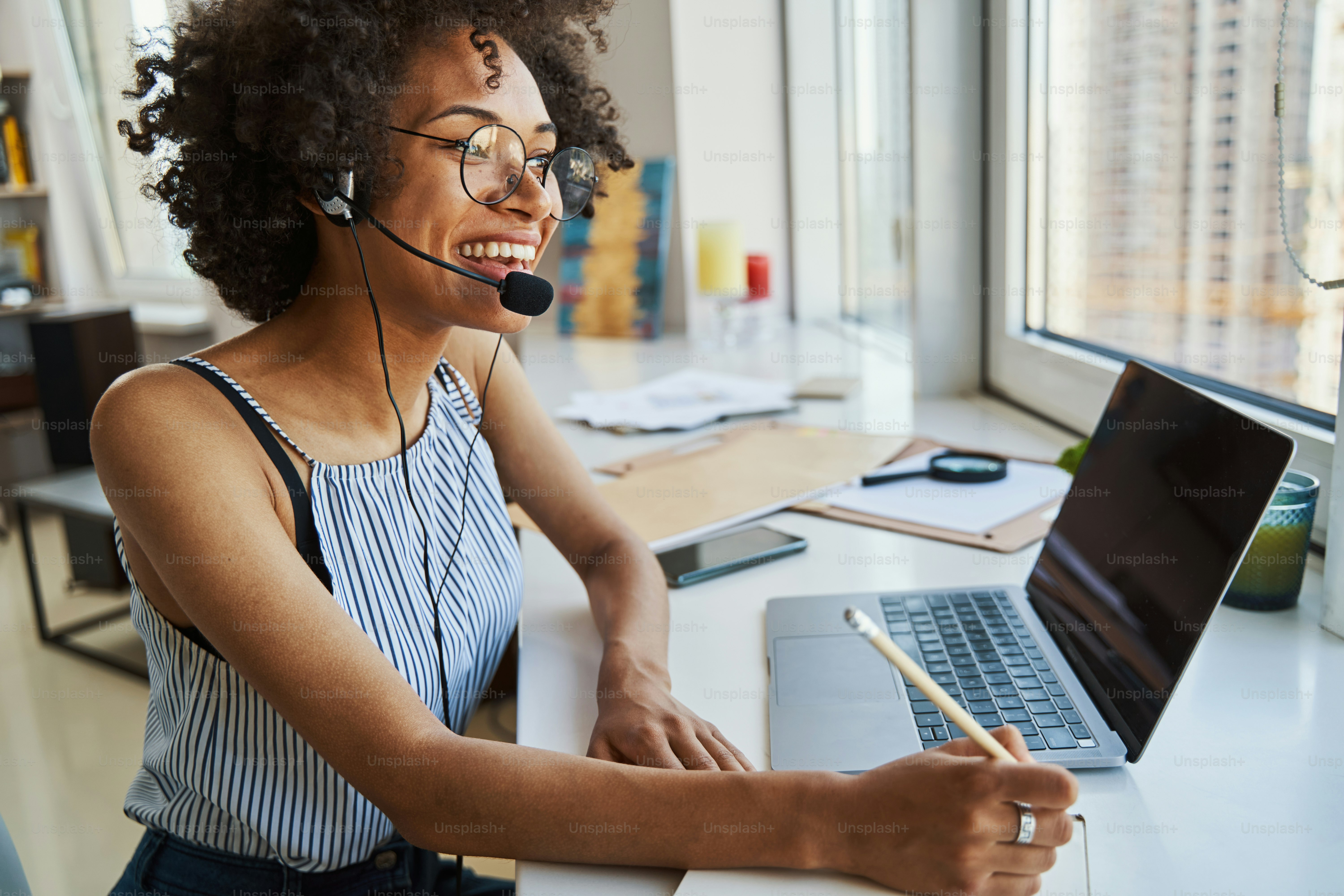 Smiling pleased female freelancer in headphones with a microphone sitting at the desk with a pencil in one hand