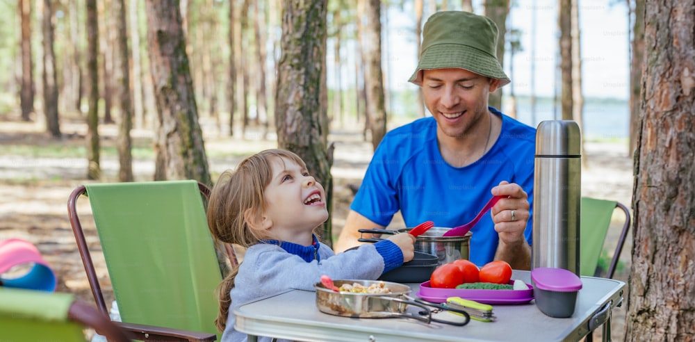 Young man father with daughter having lunch outdoor. Family holiday. Camping in the forest.