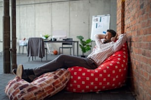 A cheerful young businessman with headphones lying on bean bag in office, taking a break and relaxing.