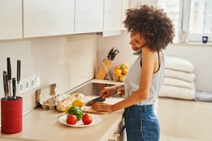Smiling woman with a smartphone pressed to her ear and a knife in her hand standing at the kitchen countertop