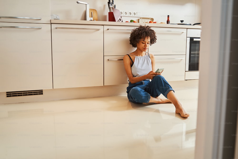 Donna di bell'aspetto seria concentrata seduta a piedi nudi sul pavimento della cucina che guarda lo schermo del suo smartphone