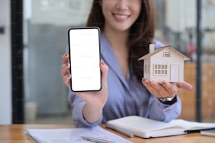 Immobilienmaklerin zeigt Smartphone mit leerem Bildschirm und Hausmodell.