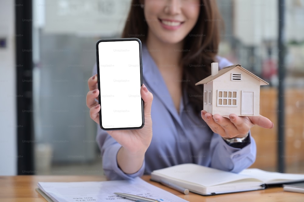 Mujer agente de bienes raíces que muestra un teléfono inteligente con la pantalla vacía y el modelo de la casa de espera.