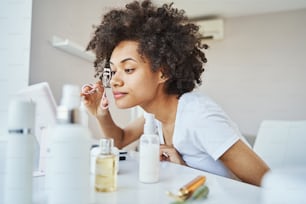 Mujer afroamericana tranquila y concentrada rizando sus pestañas con una herramienta cosmética frente al espejo