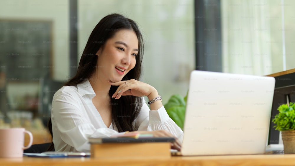 Attractive business female boss working on laptop computer in office, hand on chin, pleased with her work