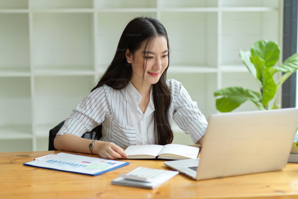 Image of young pleased happy businesswoman working on laptop computer in modern office, financial report, notebook on desk