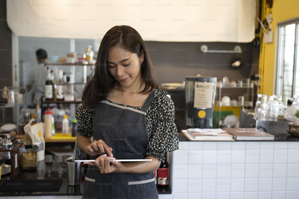 Asian woman entrepreneur standing at the counter of her cafe and using digital tablet.