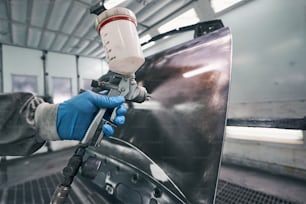 Man using spray gun for coating metal automobile part with paint layer in spraying room