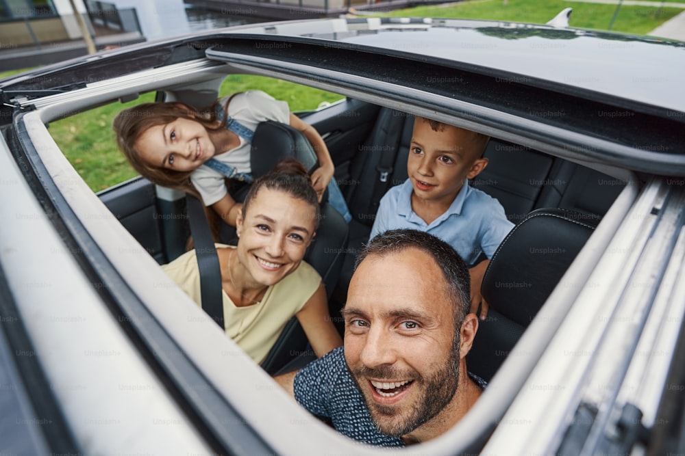 Homem contente sorrindo e olhando através do teto solar enquanto está sentado em um carro com sua amada esposa e dois filhos