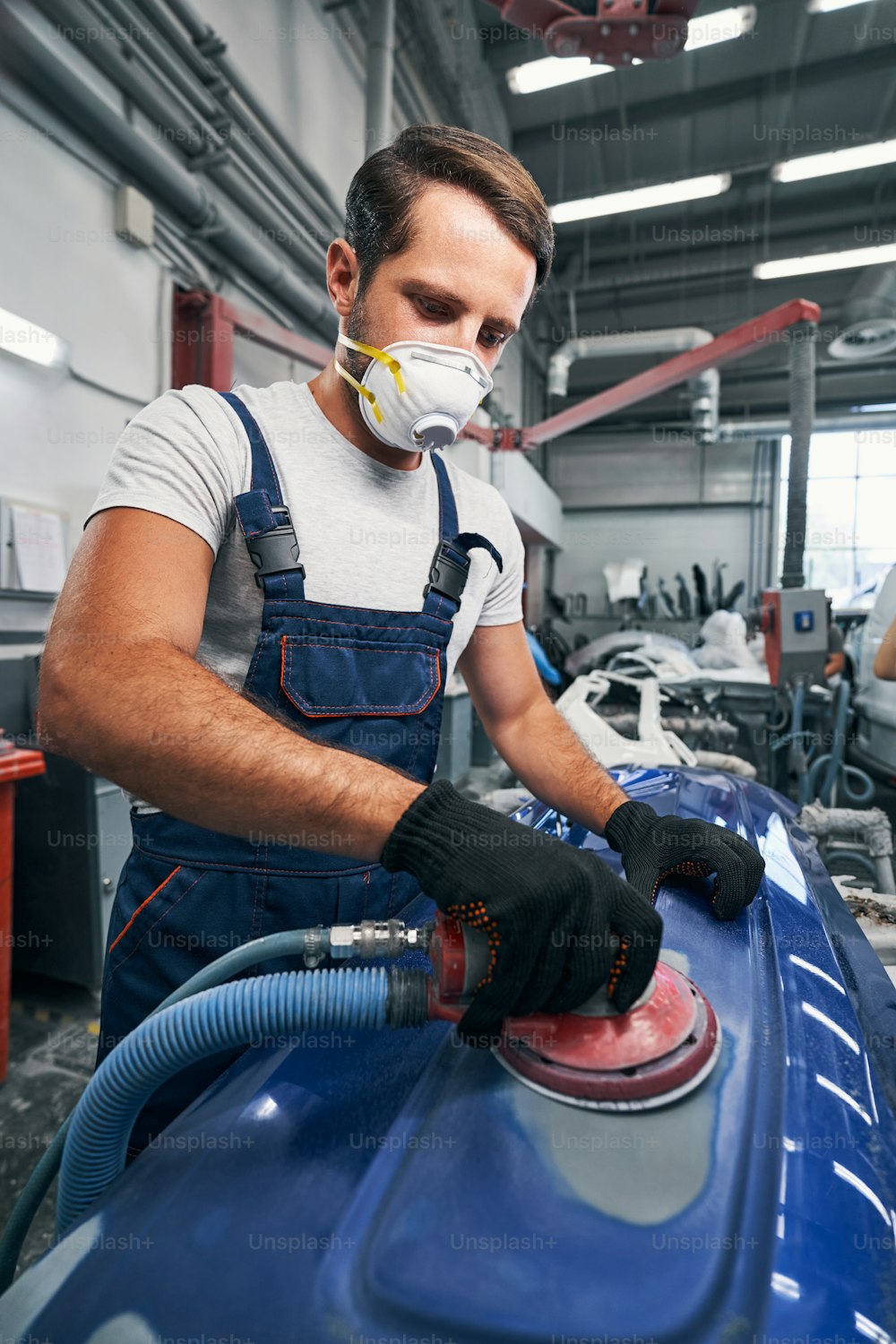 Automotive technician wearing blue overalls and breathing mask grinding part of automobile with sander in workshop