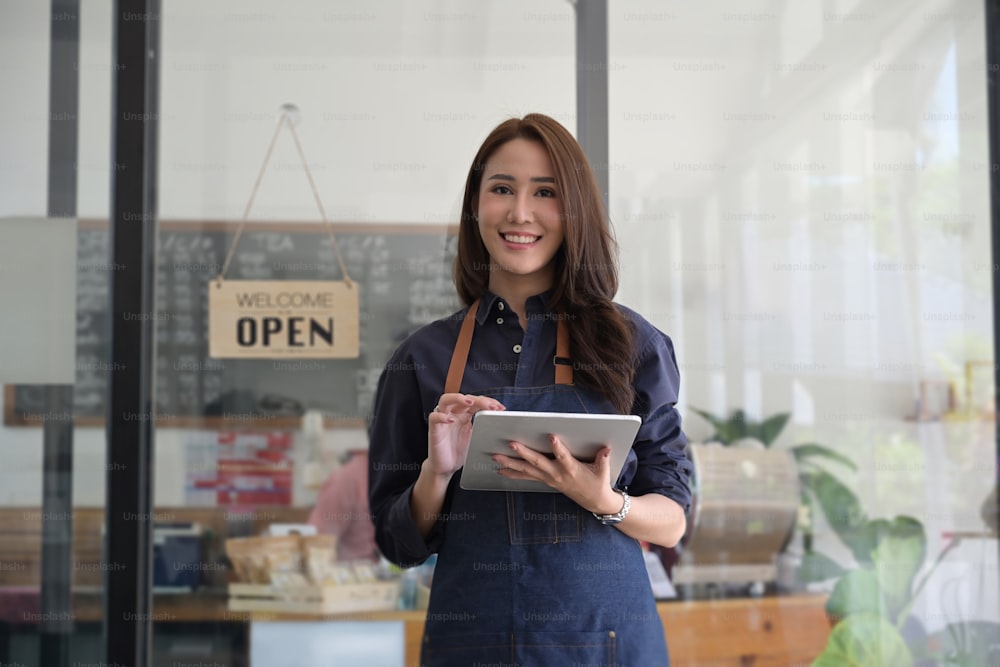 エプロン姿の若い女性起業家が、モダンなカフェに立ちながらデジタルタブレットを使用している。