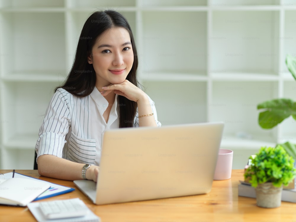 Beautiful asian businesswoman positive smile to camera, hand on chin, sitting in modern office laptop on working desk