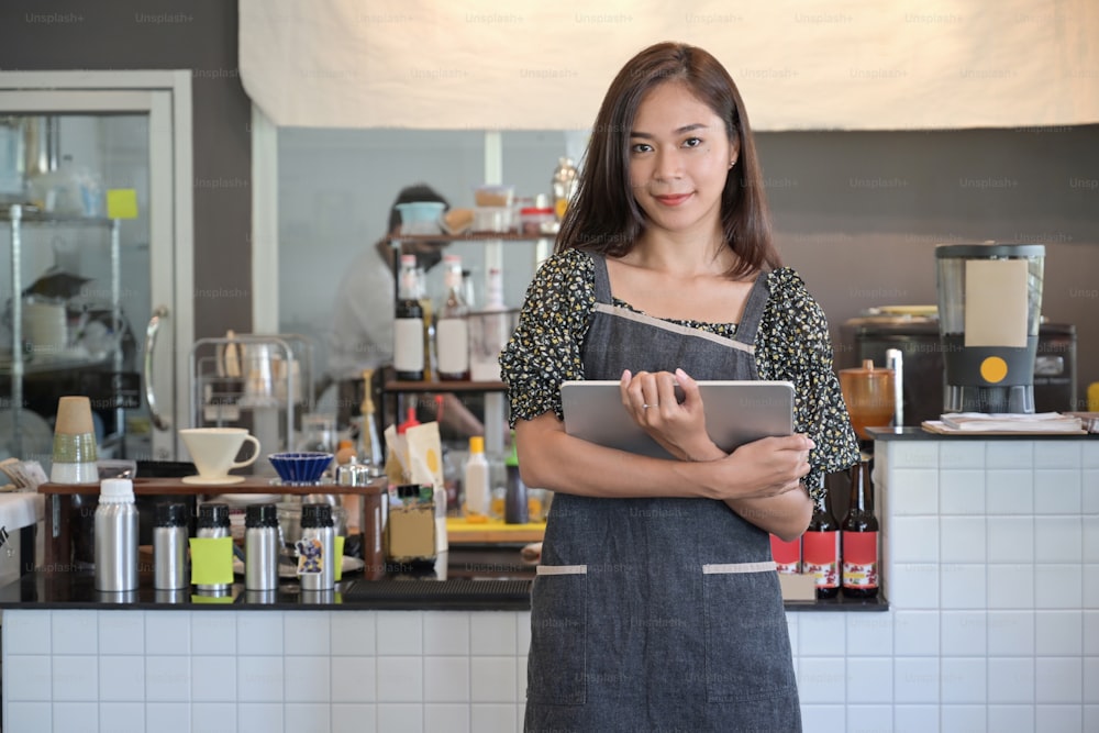 Beautiful woman small business owner holding digital tablet and standing in her coffee shop.