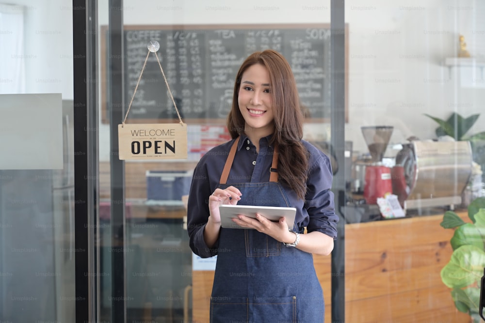 그녀의 커피숍에 서 있는 동안 디지털 태블릿을 들고 있는 친절한 여성 기업가의 초상화.