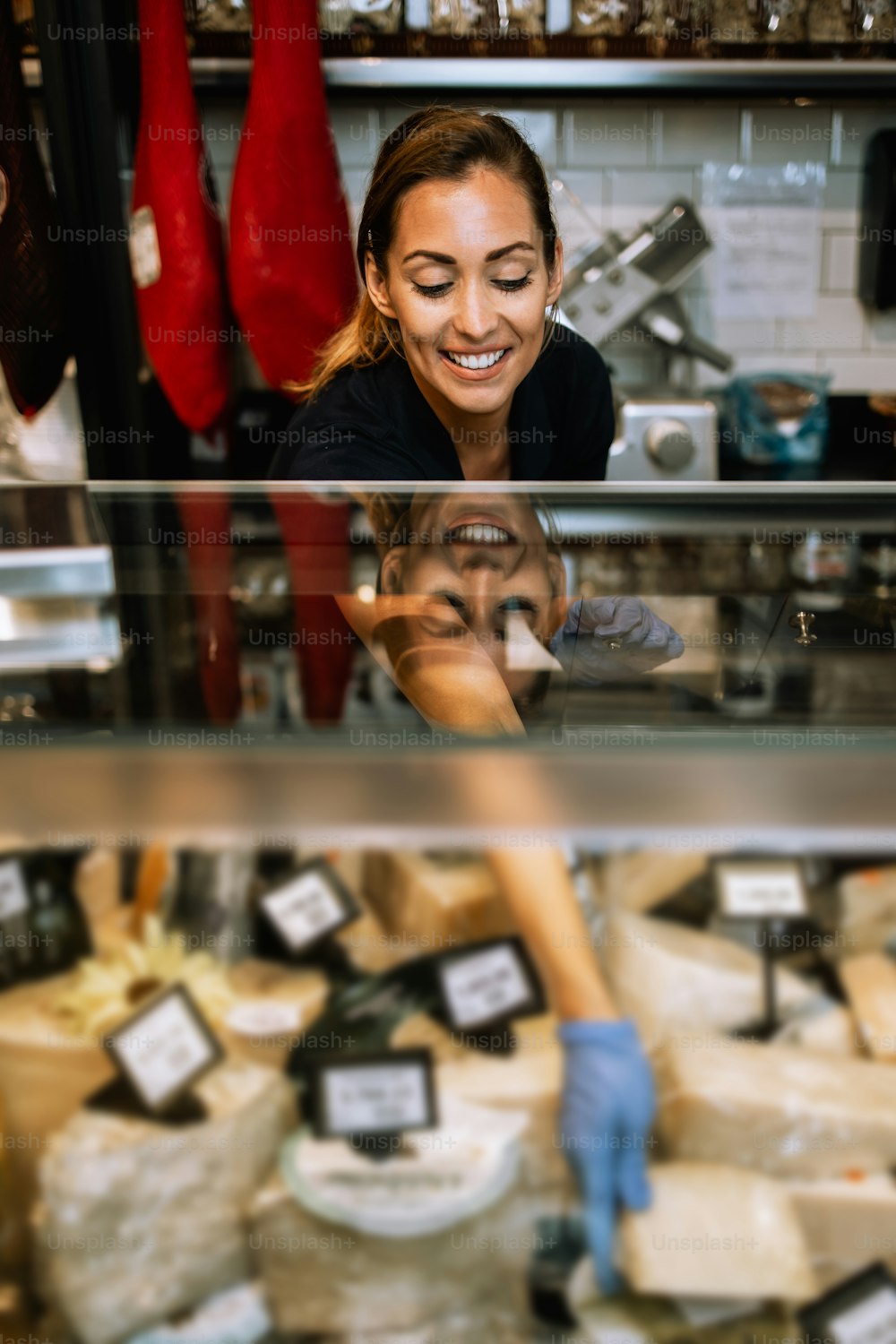 Beautiful and happy female seller working in supermarket or grocery store and selling cured meat products and cheeses.