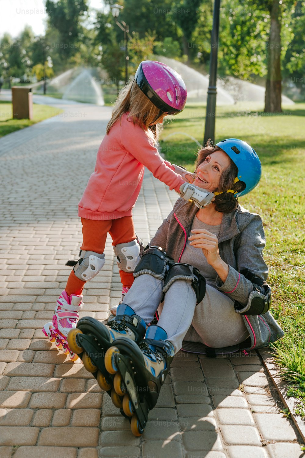 Kleines süßes Mädchen, das ihrer Großmutter hilft, sich auf das Rollschuhlaufen vorzubereiten und einen Helm im Freien trägt.