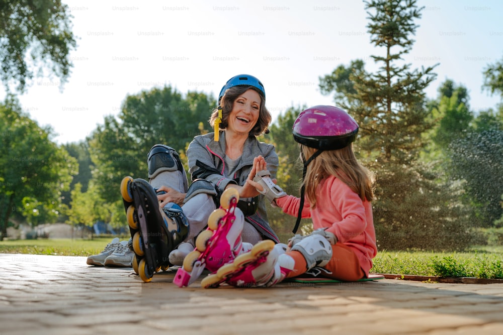 Active happy grandmother and granddaughter giving high five preparing for roller skating sitting in the park. Selective focus on the woman.