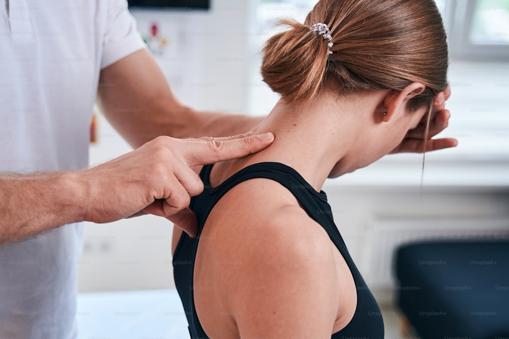 Close up back view portrait of redheaded pretty lady in black sports bra getting professional acupressure session on her neck in rehabilitation clinic