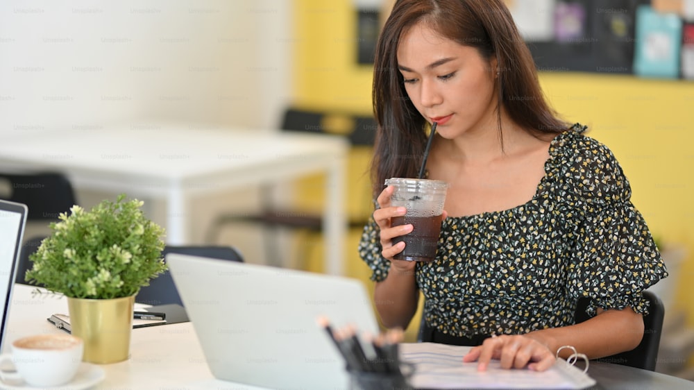 カフェで働く魅力的な女性フリーランサー、コーヒーショップでラップトップコンピューターを使用中にコーヒーを飲む