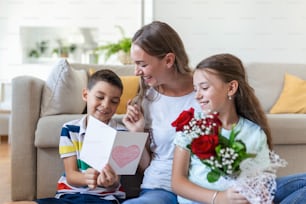 Junge Mutter mit einem Rosenstrauß lacht, umarmt ihren Sohn, und ein aufmerksames Mädchen mit einer Karte gratuliert Mama während der Weihnachtsfeier in der Küche zu Hause