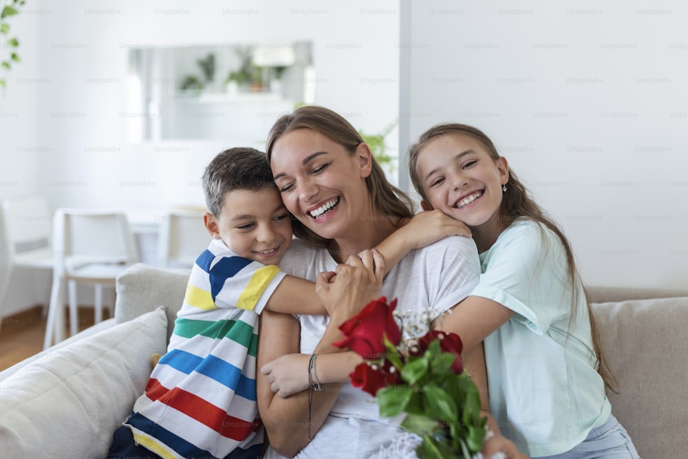 バラの花束を持った若い母親が息子を抱きしめて笑い、カードを持った元気な女の子が自宅のキッチンで休日のお祝いをしているお母さんを祝福します