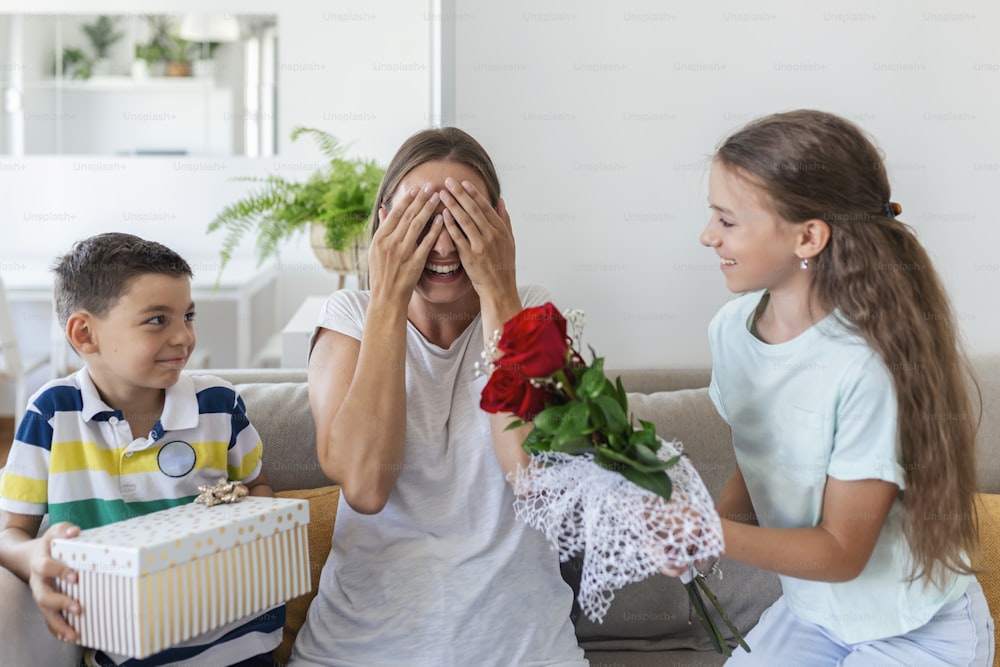 Cheerful little girl with bouquet of roses flowers and youngest brother with gift box smiling and congratulating happy mom on mother day at home. Happy Mothers Day!