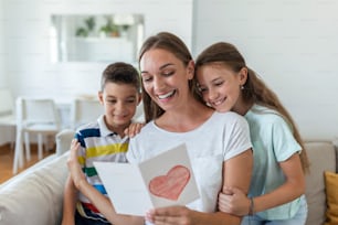 Fröhliche junge Mutter und kleine Kinder sitzen auf dem Sofa und umarmen sich, während sie Wünsche und Glückwünsche mit Urlaub in präsentierter Postkarte zu Hause lesen