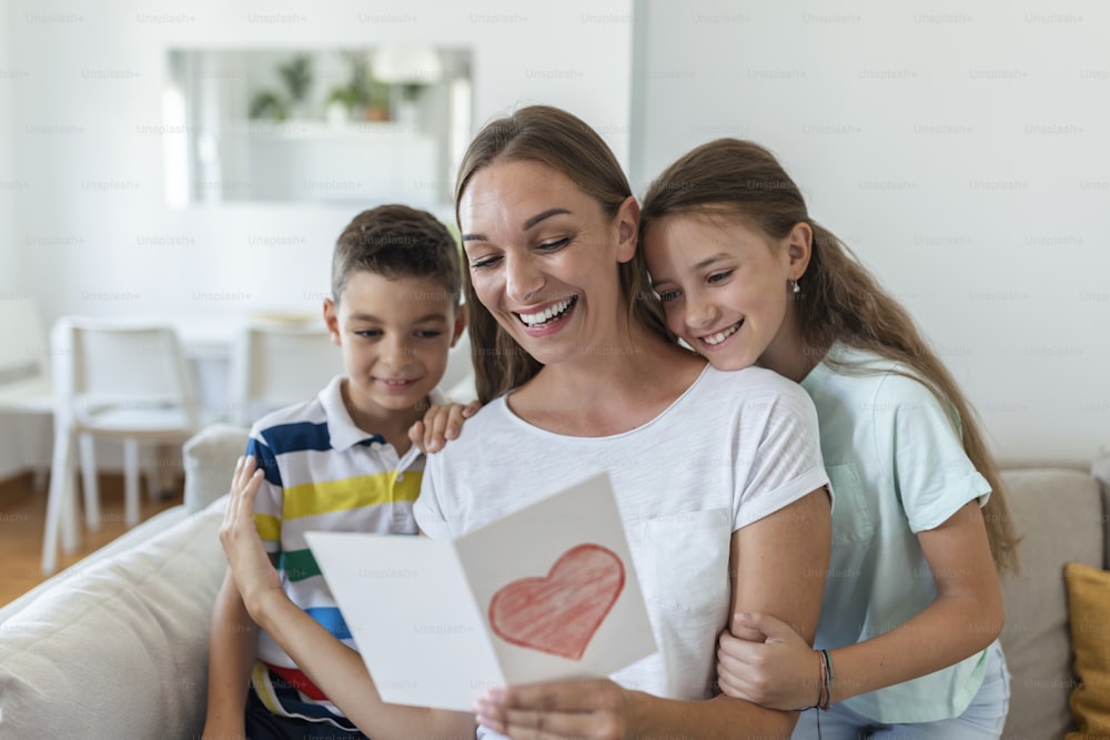 Mãe jovem alegre e crianças pequenas sentadas no sofá e abraçadas enquanto lêem desejos e parabéns com o feriado em cartão postal apresentado em casa