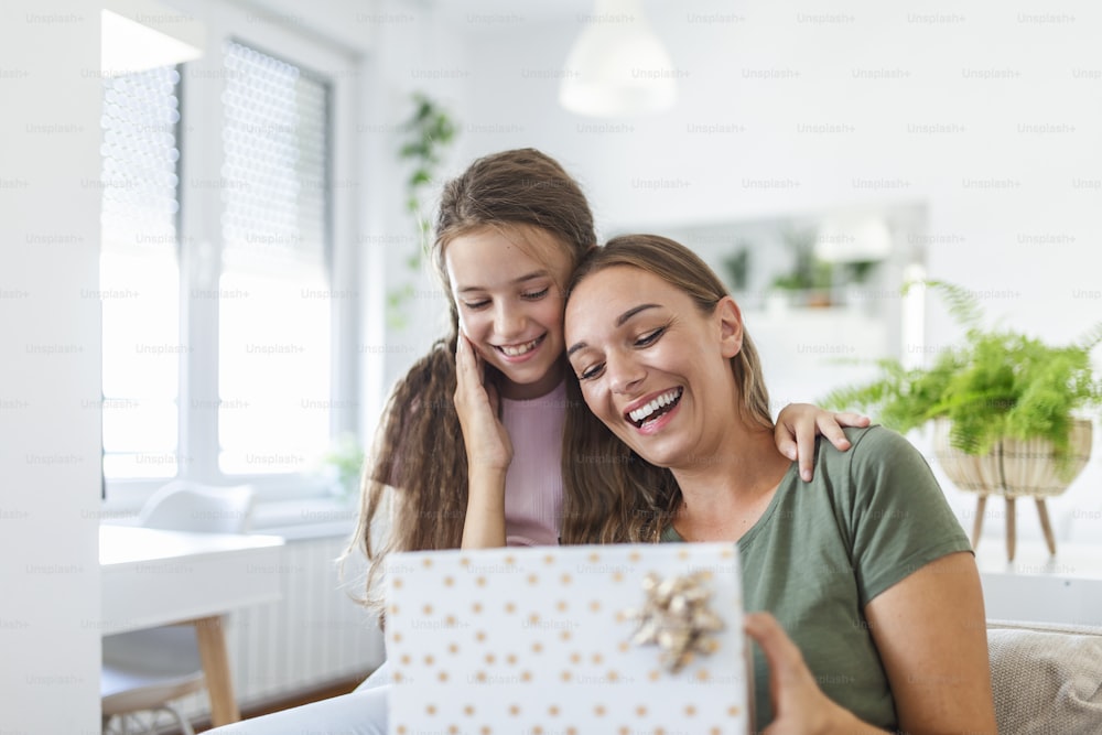 Young woman and girl at home celebrating mother's day sitting on sofa daughter hugging mother kissing cheek mom laughing joyful holding gift box