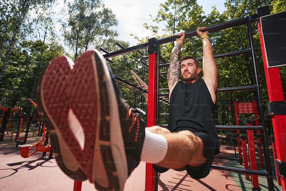 Low angle of athletic guy holding horizontal bar and lifting legs for exercising core muscles on sportsground
