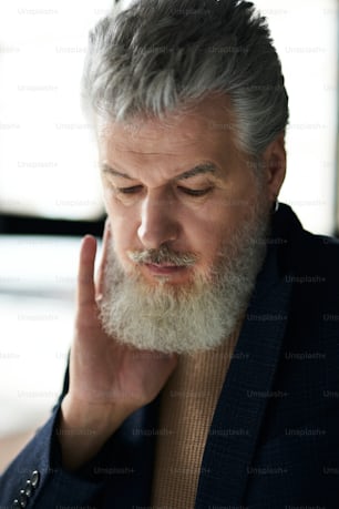 Face closeup of stylish pensive grey haired middle aged man looking down, touching his beard while posing indoors. Lifestyle, people concept