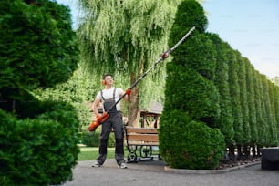 Full length portrait of strong male gardener using electric trimmer for shaping overgrown hedge outdoors. Concept of landscaping and seasonal work.