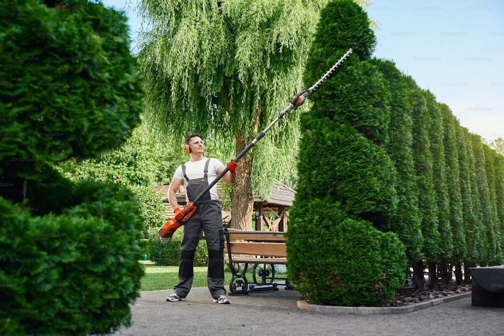 屋外で生い茂った生け垣を形作るための電気トリマーを使用した強い男性の庭師の全身の肖像画。造園と季節労働のコンセプト。