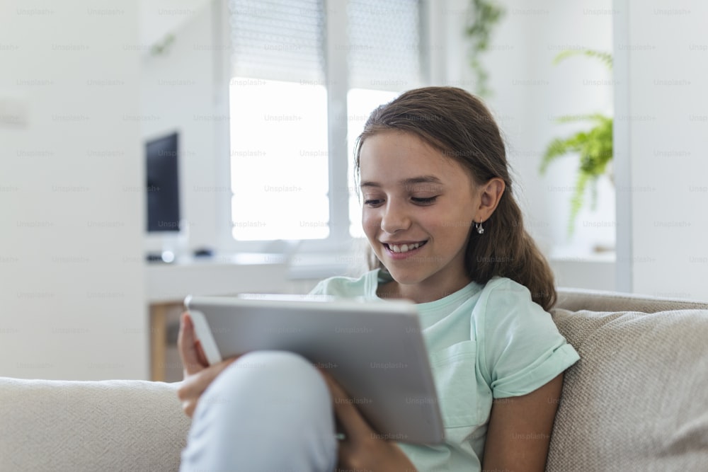 Mignon petit sourire de fille heureuse assis sur le canapé à l’aide d’un pad de tablette numérique à la recherche dans le salon à la maison. concept d’activité familiale.