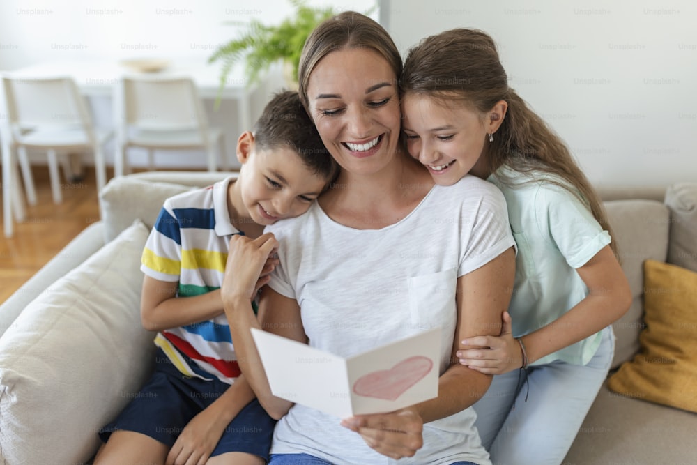 Joyful young mother and little children sitting on sofa and embracing while reading wishes and congratulations with holiday in presented postcard at home