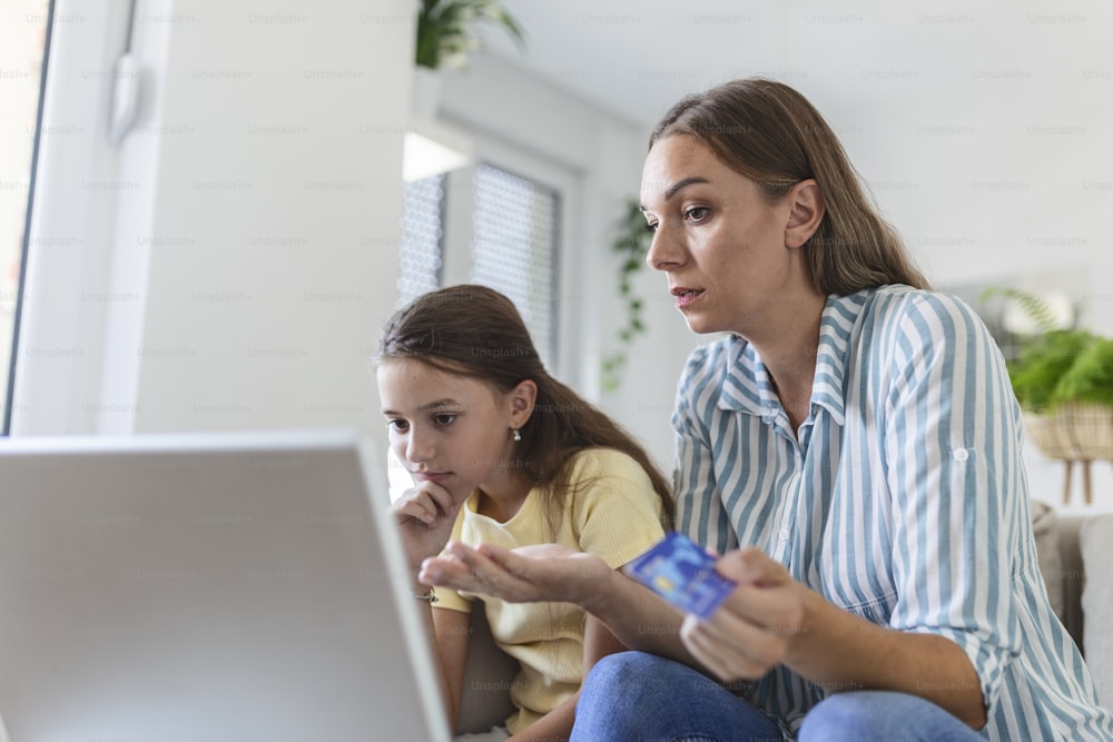 Cute small daughter rejoicing while smiling mother in casual clothes focusing on screen and using laptop for online shopping in light modern apartment