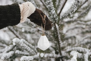 Mano en guante acogedor decorando ramas de pino con elegantes adornos blancos. Decorar árbol de navidad en nieve al aire libre. Preparación para las vacaciones de invierno en el campo. ¡Felices Fiestas!
