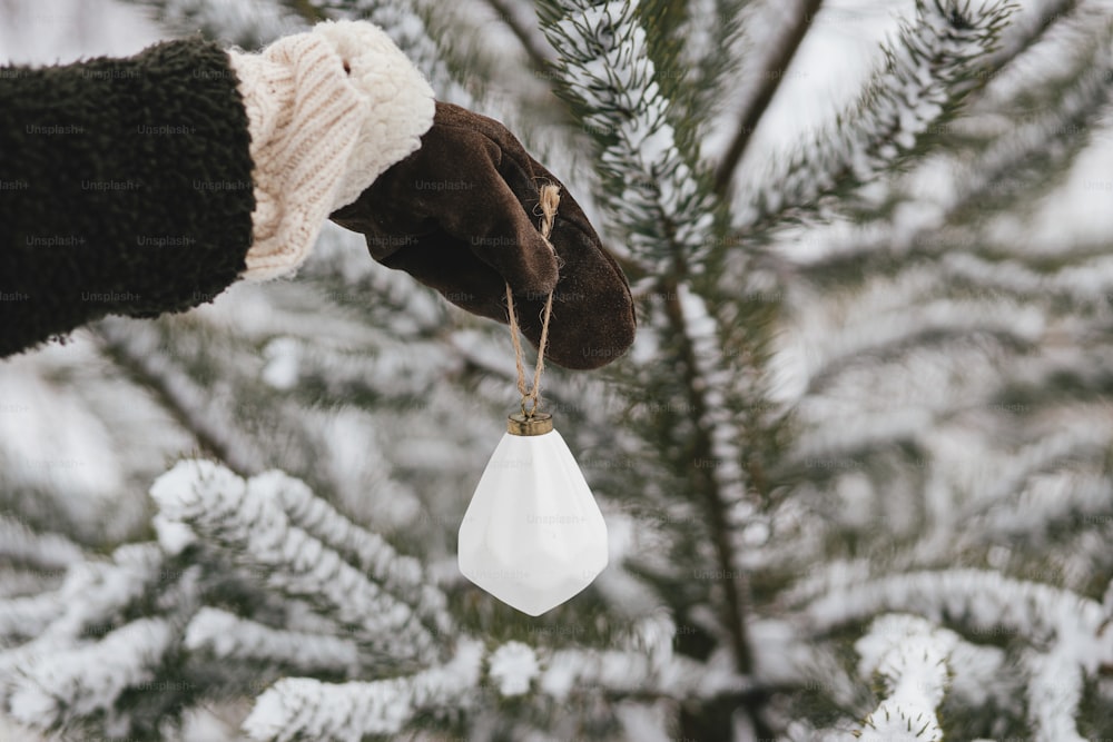 Hand in cozy glove decorating pine tree branches with stylish white ornament. Decorating christmas tree in snow outdoors. Preparation for winter holidays in countryside. Happy Holidays!