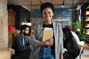 Successful smiling mixed-race businesswoman standing in creative office and looking at camera. Portrait of beautiful business woman standing in front of business team at modern agency with copy space