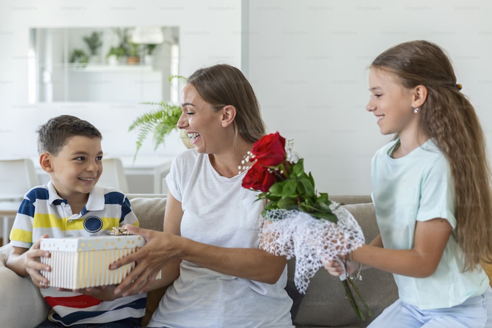 Fröhliches kleines Mädchen mit Rosenstrauß Blumen und jüngster Bruder mit Geschenkbox lächelt und gratuliert glücklicher Mama am Muttertag zu Hause. Alles Gute zum Muttertag!