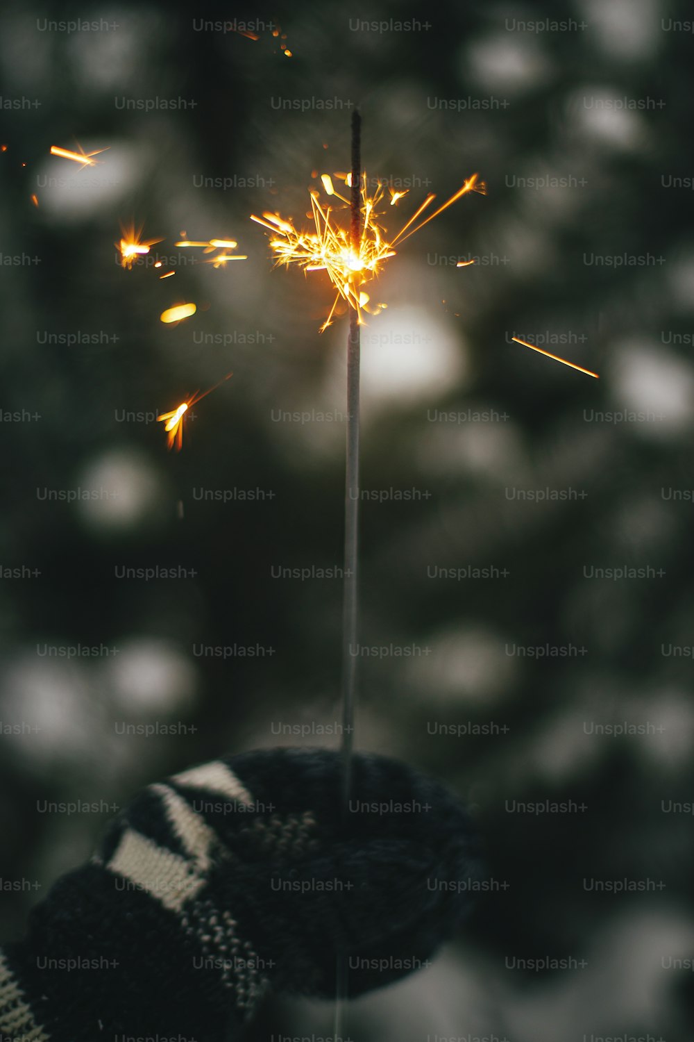 Hand in cozy mitten holding burning sparkler on background of pine tree branches in snow. Happy New Year! Atmospheric magic moment. Woman hand with glowing firework in evening.