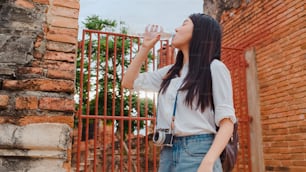 Young Asian backpacker woman blogger tourist with camera feel tired stand in front of pagoda take a rest and drink water in plastic bottle at old town , Lifestyle tourist travel holiday concept.