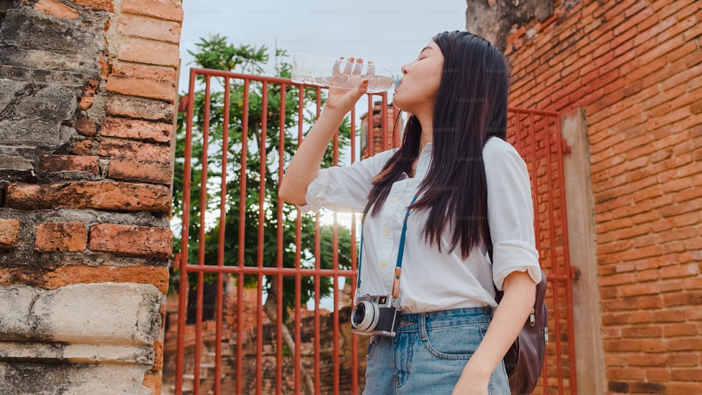 Young Asian backpacker woman blogger tourist with camera feel tired stand in front of pagoda take a rest and drink water in plastic bottle at old town , Lifestyle tourist travel holiday concept.