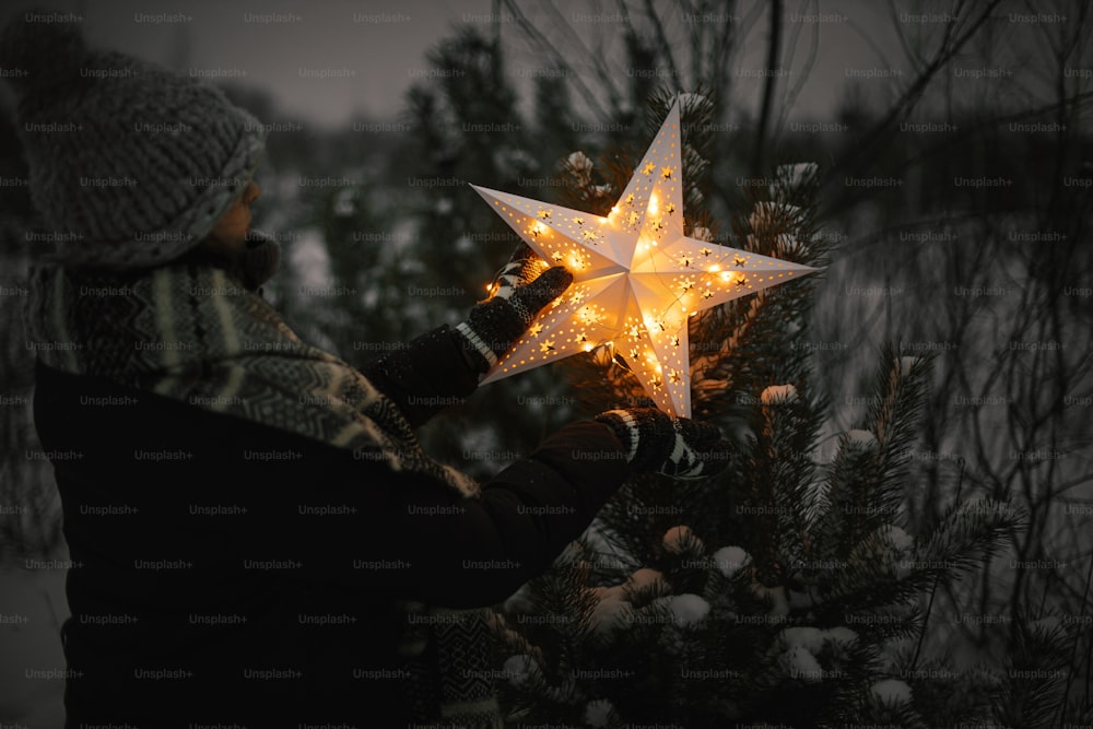 Buon Natale! Donna che mette la grande stella di Natale illuminata sul pino nel parco invernale innevato di notte. Magico periodo invernale. Elegante donna hipster che decora l'albero di Natale. Miracolo di Natale