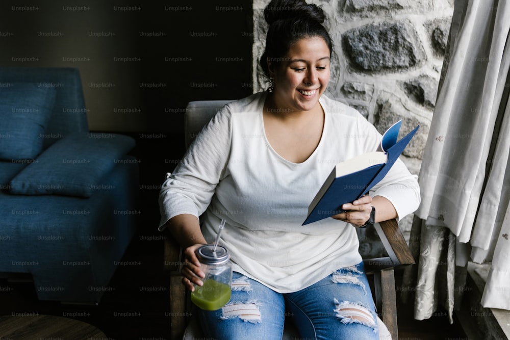 Retrato de una chica latina con curvas sosteniendo un frasco de jugo verde mientras lee un libro en casa