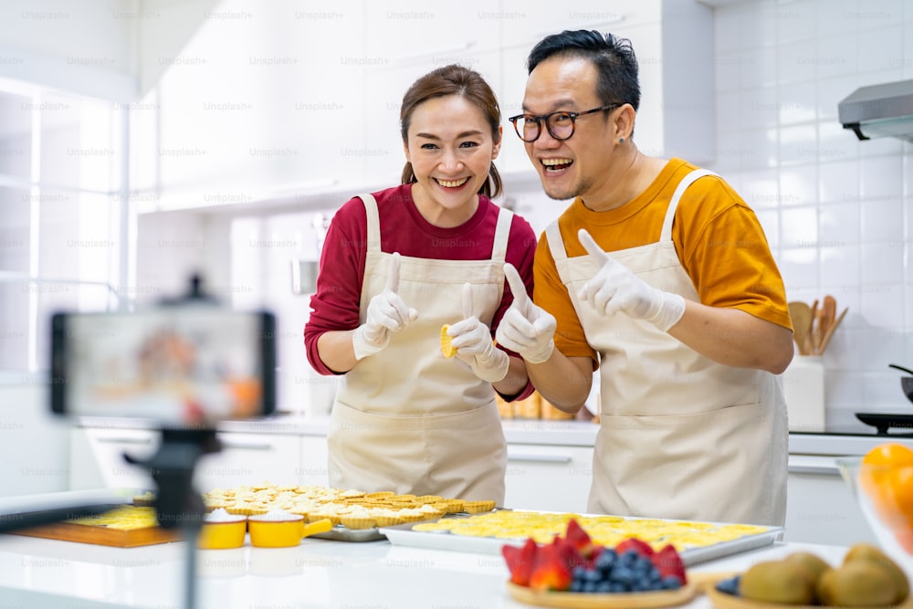 Asian couple bakery shop owner using smartphone with internet vlogging sweet dessert baking on social media together in the kitchen. Small business entrepreneur and online cooking class concept