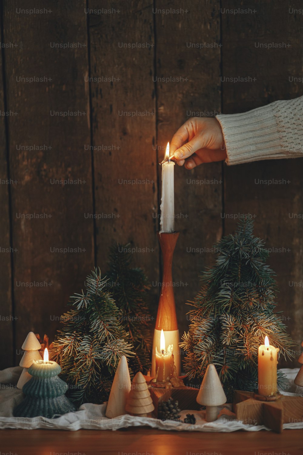 Holiday advent. Hand in cozy sweater lighting up christmas candle on rustic wooden background with pine trees and cones in evening scandinavian room.  Atmospheric moment
