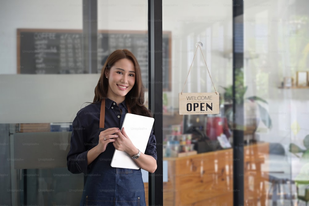 デジタルタブレットを持ち、モダンなコーヒーショップに立っている笑顔の女性ウェイトレス。
