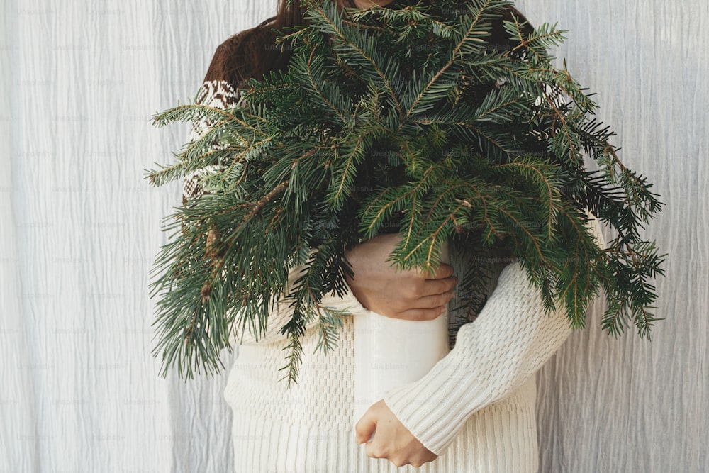 Mujer con suéter acogedor sosteniendo ramas de pino y abeto en un jarrón en una habitación rústica. Preparativos para las vacaciones de invierno. Horario de invierno atmosférico. Higiene escandinava